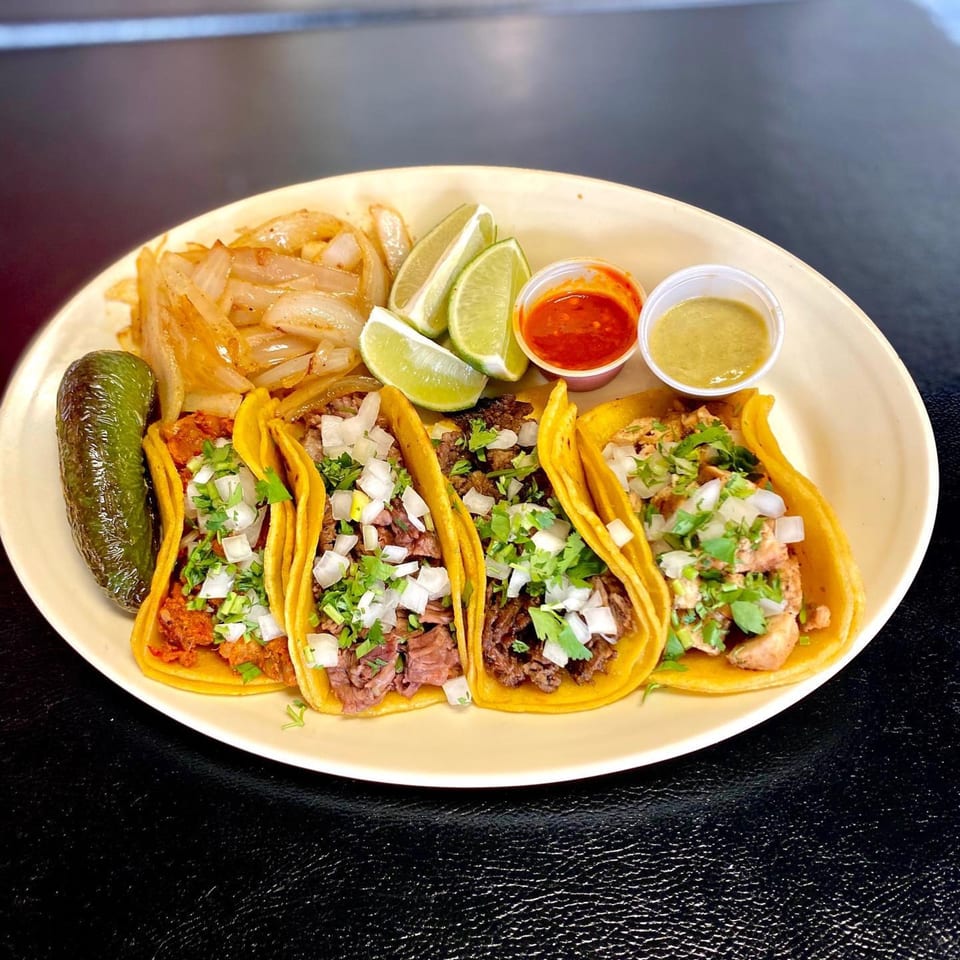 Four Dona Mary tacos with various fillings, cilantro, onions, lime, grilled onions, and sauces.