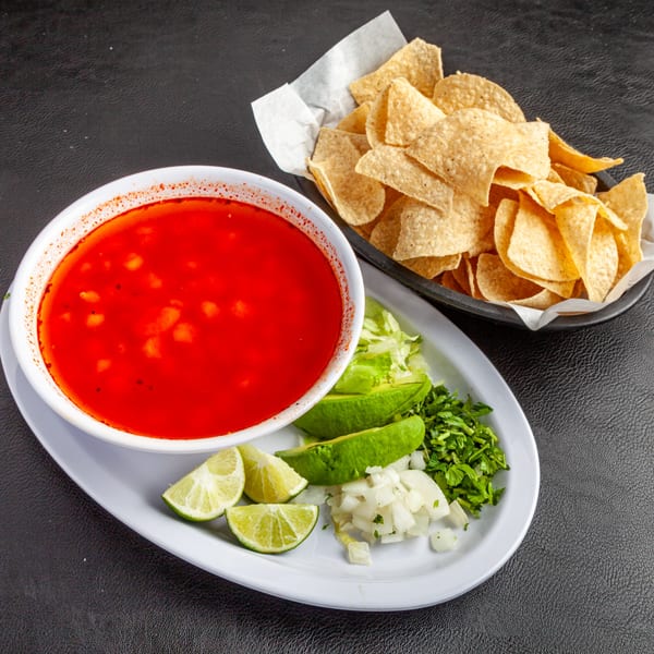 Dona Mary's red pozole soup with hominy, served with a side of tortilla chips, limes, avocado, onion, and cilantro.